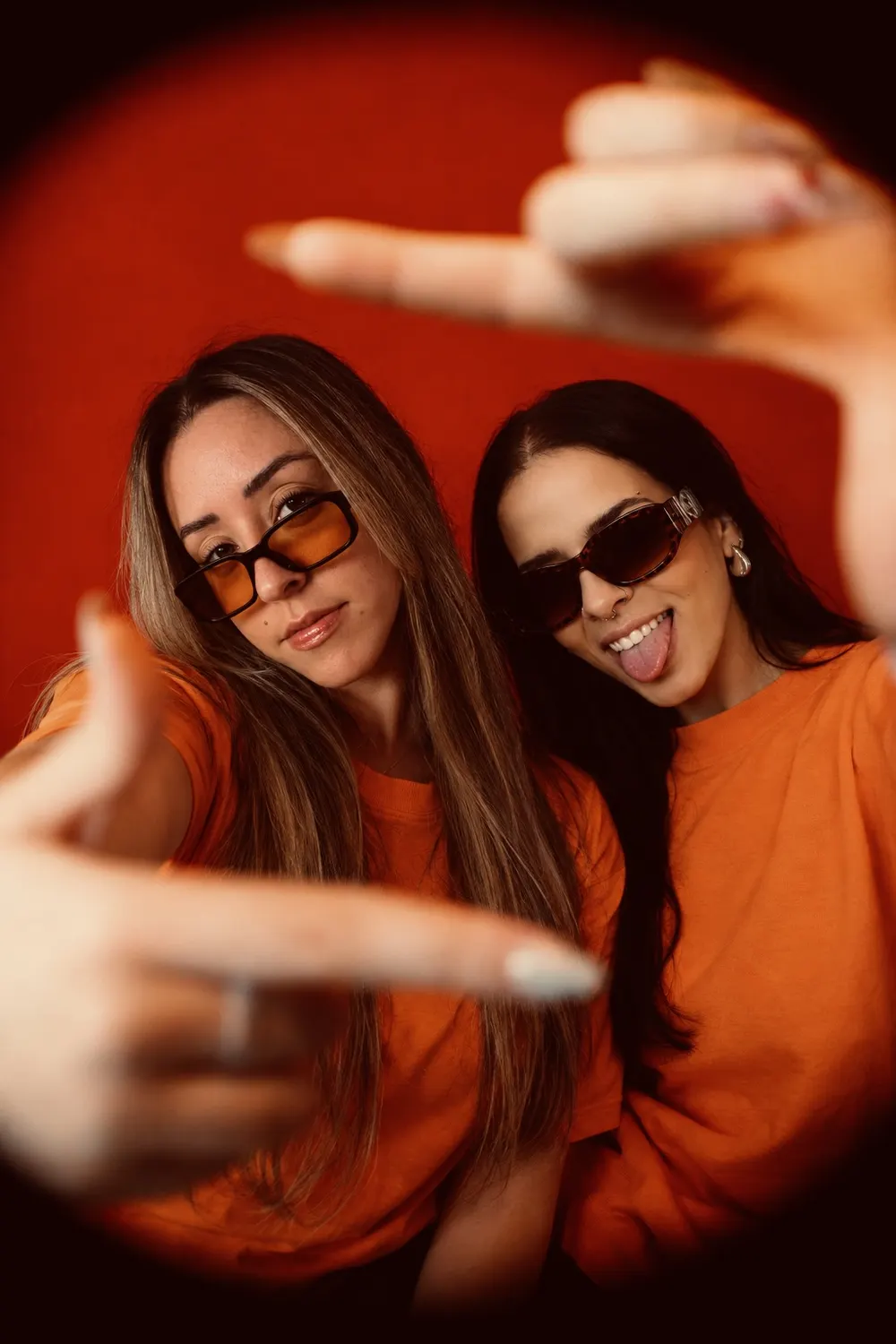 Duda e Camila enquadrando a câmera com as mãos, sobre fundo vermelho
